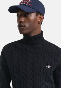 Schwarzer Pullover mit Kabelstrickmuster, Stehkragen und kleinem besticktem Logo. Kombiniert mit einer marineblauen Mütze mit roterschrift.