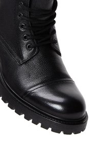 Bota de cuero negro hasta el tobillo con parte superior texturizada, puntera redonda y suela gruesa y resistente. Cuenta con cordones negros y herrajes de ojal.