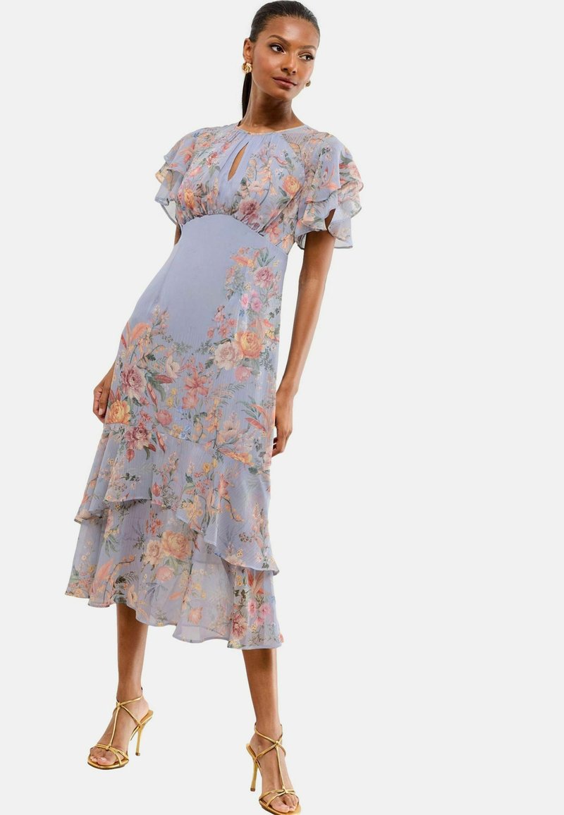 Abito midi floreale azzurro con maniche a volant e strati, caratterizzato da un collo a chiave e una silhouette fluida. Sandali dorati con strap.