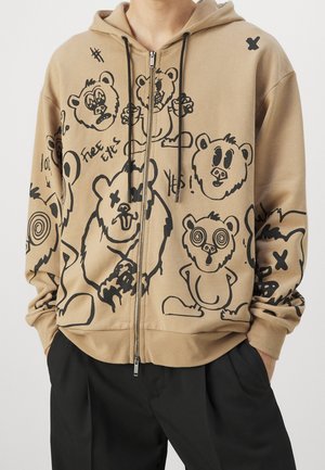 Beiger Kapuzenpullover mit Reißverschluss, schwarzen Comic-Bärengesichtern und Texten wie "Free Eyes" und "Yes!", getragen von einer Person mit Händen in den Taschen schwarzer Hosen.
