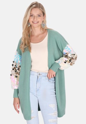 Cardigan verde chiaro con maniche decorate con paillettes; texture in maglia morbida; design aperto davanti; abbinato a una maglietta crema e jeans strappati blu chiaro.