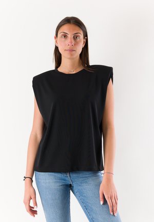 Donna con capelli castani che indossa un top nero senza maniche con spalle imbottite e jeans blu, in piedi davanti a uno sfondo bianco semplice.