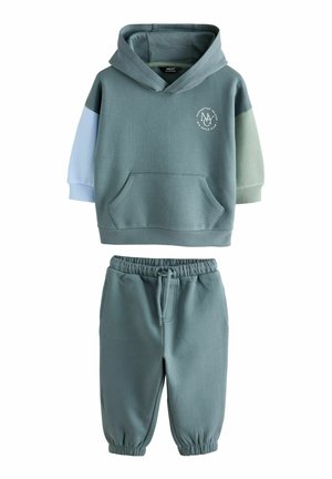 Dwuczęściowy zestaw składający się z niebieskozielonego hoodie z różnymi kolorami rękawów oraz kieszenią kangurką, połączony z niebieskozielonymi spodniami dresowymi z elastycznymi mankietami.
