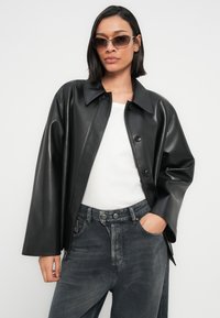 Μαύρο μπουφάν από συνθετικό δέρμα με γιακά πουκαμίσου, δύο κουμπιά και φαρδιά εφαρμογή. Φορεμένο πάνω από λευκό τοπ και συνδυασμένο με σκουρόχρωμο γκρι τζιν παντελόνι.