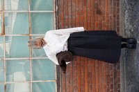 Fehér, gombos ing nagy gallérral, fekete textúrájú szoknya, barna kabát és fekete bokacsizma; tégla fal előtt.