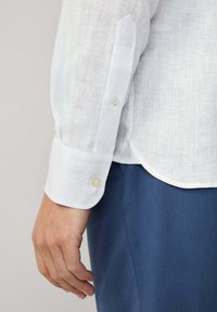 Hand aan de zijkant, gekleed in een wit linnen overhemd met lange mouwen en knoopmanchetten, en blauwe broek, lichte neutrale achtergrond.