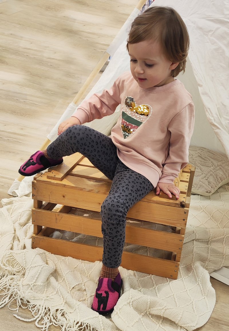Lichtroze sweatshirt met een pailletten hart ontwerp, grijze luipaardprint leggings en roze slippers met zwarte accenten. Zittend op een houten krat.