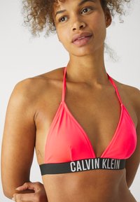 Top de bikini triangular en tela rosa brillante con tirantes finos y una banda negra que presenta "CALVIN KLEIN" en texto blanco. Textura suave.