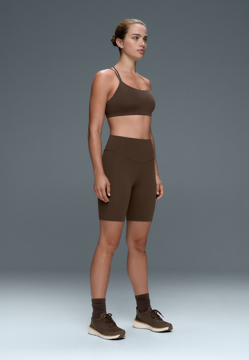 Young woman standing wearing matching brown sports bra, high-waisted bike shorts, brown socks, and brown sneakers against gray background.