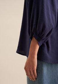 Granatowa bluzka z bufiastymi rękawami, z detalem guzika przy mankiecie, wykonana z lekkiej, teksturowanej tkaniny. Luźny krój i styl.