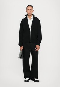 Black ribbed zip-up cardigan with a high collar, worn over a white shirt. Paired with loose black trousers and black shoes. Holding a grey bag.