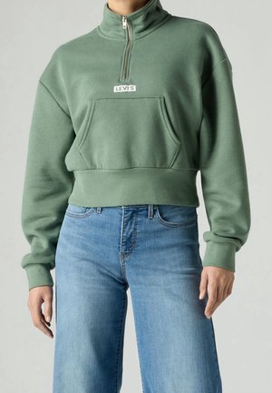 Person iført en grøn Levi's quarter-zip sweatshirt med kængurulomme og lyseblå højtaljede jeans mod en ensfarvet baggrund.