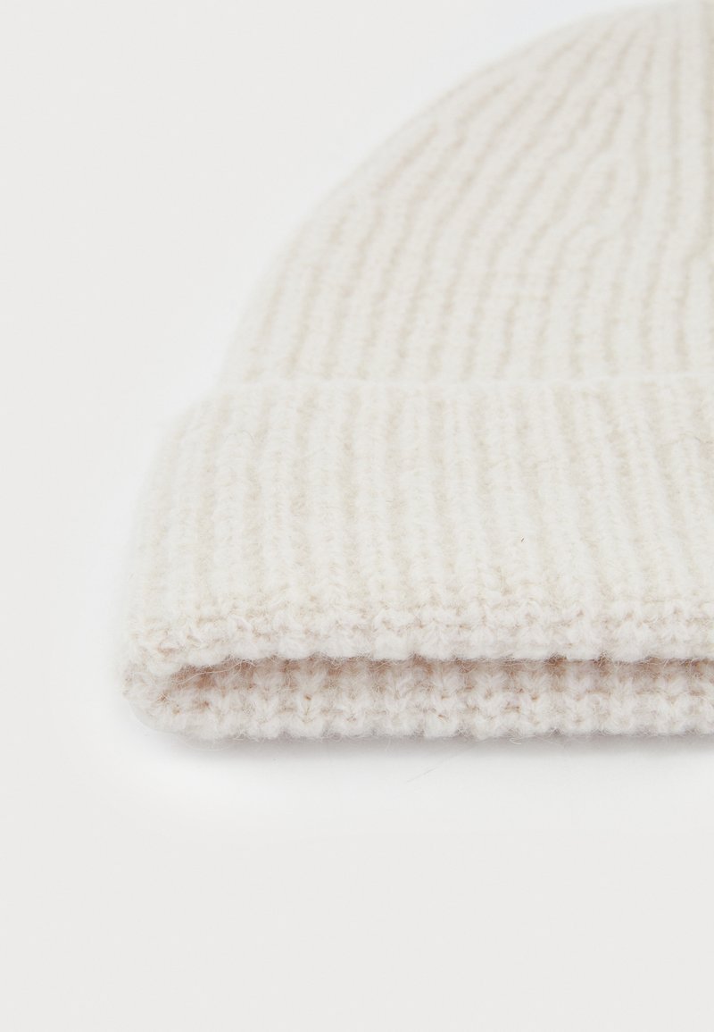 Strikket hue i lys beige uld, med ribbete tekstur og en foldet kant i bunden, der viser blødt og varmt materiale.