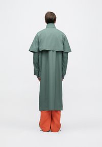 Dlhý zelený kabát so zvonovým strihom a vysokým golierom, nosený na jasne oranžových nohaviciach, s asymetrickým vrstveným chrbtom.