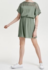 Vestido verde de manga corta con diseño de encaje en el escote, cintura elástica y una silueta fluida; combinado con zapatillas blancas.