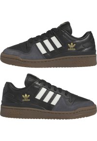 Schwarze und graue Adidas-Sneaker mit weißen Streifen, goldenem Logo, braunen Sohlen und schwarzen Schnürsenkeln, aus seitlicher und schräger Perspektive gezeigt.