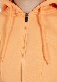 Hoodie orange à fermeture éclair en tissu texturé, avec une fermeture éclair noire, des cordons de serrage et des accents cousus le long des coutures.