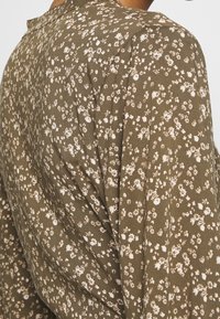 Tessuto verde oliva con un piccolo motivo floreale bianco, drappeggiato con una texture morbida e rilassata e una leggera lucentezza.