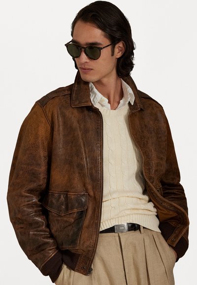 Chaqueta bomber de cuero marrón con una textura desgastada, que presenta puños acanalados, cierre frontal y bolsillos laterales. Llevada sobre un suéter de punto trenzado.