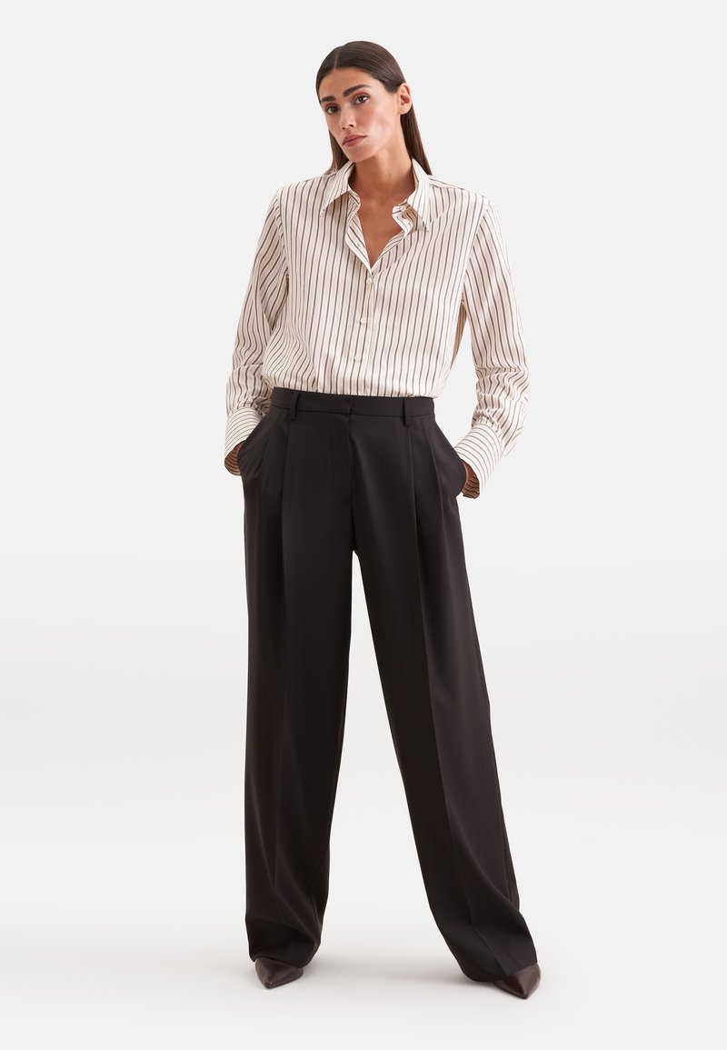 Femme aux longs cheveux foncés debout, portant une chemise crème rayée à boutons, rentrée dans un pantalon noir taille haute à jambes larges, les mains dans les poches.
