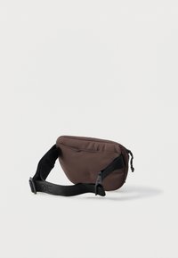 Ruskean kankaan vyölaukku, jossa on vetoketjukiinnitys, pehmustettu muotoilu ja musta säädettävä hihna, jossa on logot yksityiskohtina. Sileä pinta ja kompakti muoto.