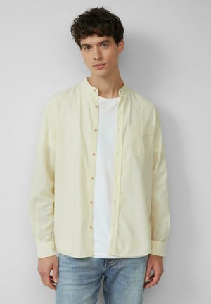 Chemise à boutons jaune clair en tissu doux et texturé, avec un col montant et une poche poitrine, portée par-dessus un t-shirt blanc.