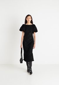 Črna, prilegajoča midi obleka s pufastimi rokavi, teksturirano blago, kvadratnim izrezom in subtilnim vzorcem. Nesha s črnimi škornji do kolen in torbico.