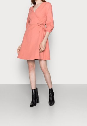 Kvinde iført en knælængde koralfarvet slå-om-kjole med trekvart puffede ærmer og sorte ankelstøvler med hæl på ensfarvet baggrund.