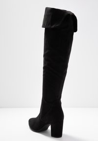 Korkeat mustat mokkanahkabootsit, joissa on taitettu varsi, pyöreä kärki ja tukeva lohkoiluhe, valkoista taustaa vasten.