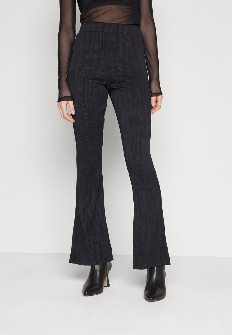 Pantalon flare noir texturé taille haute, associé à des bottines noires à talons et un haut transparent à manches longues.