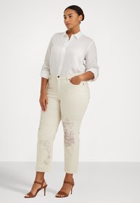 Camisa de lino blanca con botones, combinada con pantalones crema con diseños impresos en púrpura y una silueta ajustada. Sandalias marrones de tacón.