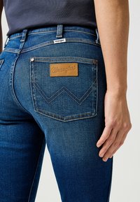 Jeans de mezclilla azul con un diseño ajustado, que presentan una etiqueta de cuero marrón con el logo "Wrangler" y bolsillos traseros con costuras decorativas.