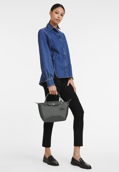 Gray handbag with a textured black handle, featuring a green zipper accent. Model wears a blue denim shirt, black pants, and loafers.