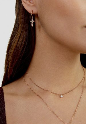 Donna che indossa un orecchino a cerchio d'oro con una piccola croce di cristallo e due delicati collane d'oro, una con un ciondolo di cristallo singolo.