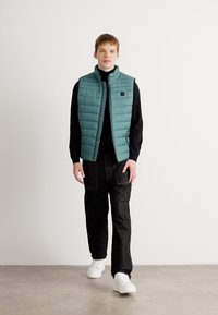 Gilet rembourré teal avec un col haut, porté par-dessus un col roulé noir, associé à un pantalon cargo noir et des baskets blanches. Design minimaliste.