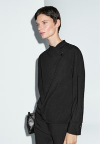 Top nero con collo alto, caratterizzato da un tessuto drappeggiato e un dettaglio con bottone laterale. Abbinato a pantaloni neri e una piccola borsa nera.