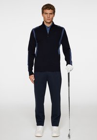 Marinblå tröja med fjärdedels dragkedja, med ljusblå detaljer på axlarna. Kombinerad med mörka byxor och vita skor, håller en golfklubba.