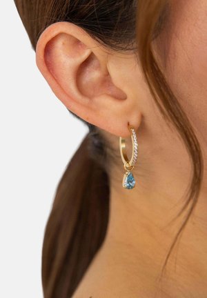 Goldener Kreolen-Ohrring mit kleinen klaren Steinen und einem hängenden tropfenförmigen blauen Edelstein am Ohr einer Frau mit braunen Haaren.