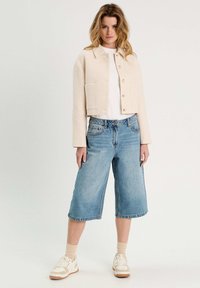 Cremfarbener, strukturierter, gekürzter Blazer, weißes T-Shirt, hellblaue, weite Denim-Shorts und weiße Sneakers mit beigen Sohlen, stehend vor einem schlichten Hintergrund.