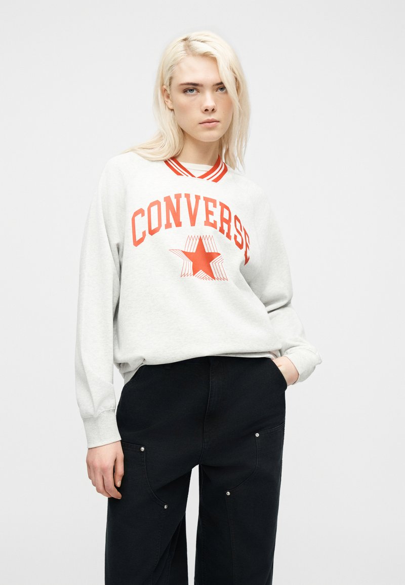 Junge Frau mit blonden Haaren, die ein hellgraues Converse-Sweatshirt mit rotem Sternlogo und schwarze Hose trägt, stehend vor weißem Hintergrund.