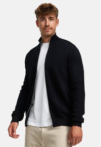 Cárdigan negro con cremallera y un patrón de zigzag texturizado, con puños y collar acanalados, llevado sobre una camiseta blanca y pantalones de colores claros.