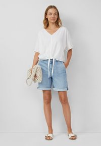 Weißes, lockeres V-Ausschnitt-Shirt mit einer Frontbindung. Hellblaue Denim-Shorts mit umgeschlagenen Säumen. Bequeme Sandalen und eine geflochtene Tasche.