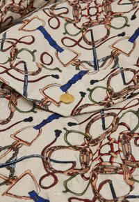 Tuvumā redzams krēmu audums ar jāšanas iedvesmotu rakstu, kas satur siksnas, virves un zeltītu pogu ar ūdens pilēm uz virsmas.