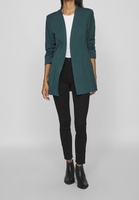 Tyrkysový blazer se strukturovaným designem, otevřeným předním dílem a dlouhými rukávy, doplněný bílým topem a černými skinny jeans, zakončený černými kotníkovými botami.