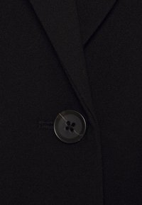Gros plan d'un blazer en tissu noir avec un seul bouton noir à quatre trous fixé sur le devant.