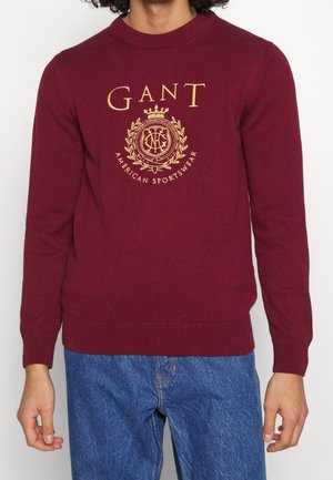 Homme portant un pull bordeaux avec un logo et un emblème dorés GANT, associé à un jean en denim bleu, debout devant un fond clair.