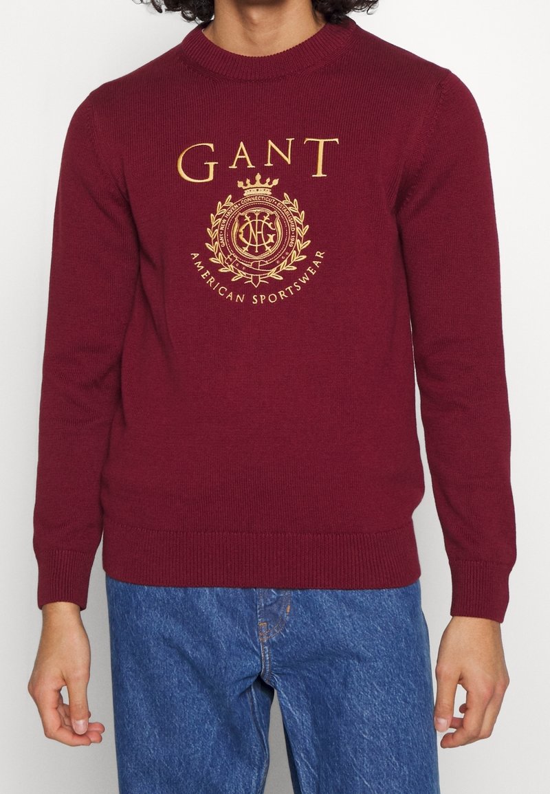 Homme portant un pull bordeaux avec un logo et un emblème dorés GANT, associé à un jean en denim bleu, debout devant un fond clair.