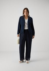 Navy blazer over a white textured top, paired with navy pleated wide-leg pants. Accessories include a pearl beaded bag and white sandals.