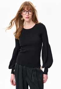 Młoda kobieta z długimi włosami i okularami, ubrana w czarny prążkowany sweter z bufkami oraz luźne czarne spodnie na białym tle.