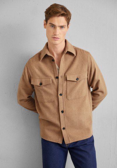 Braune Wollhemdjacke mit Knopfleiste, zwei Brusttaschen und klassischem Kragen, kombiniert mit dunkelblauen Jeans.